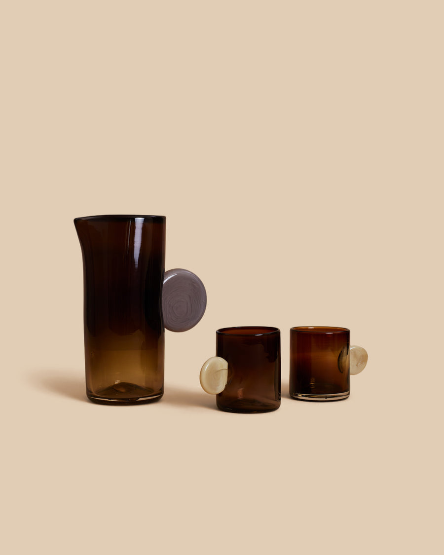 Dot Carafe and Dot Cup Set by Sticky Glass in amber and cream, a tall smoky brown carafe with a lilac disc handle beside two amber cups with cream discs