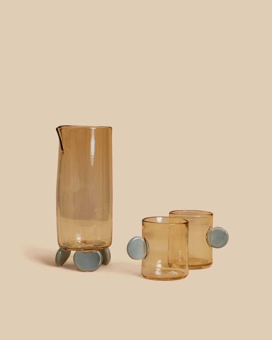 Dot Carafe and Dot Cup Set by Sticky Glass in whiskey and grey, a pale amber carafe standing on blue-grey glass feet with two matching cups