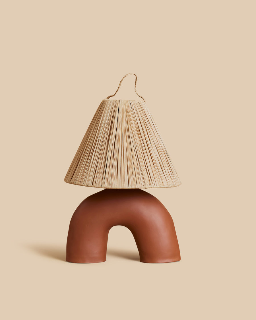 Volta Lamp by Marta Bonilla, a terracotta table lamp with an arched half-circle base under a pleated raffia shade