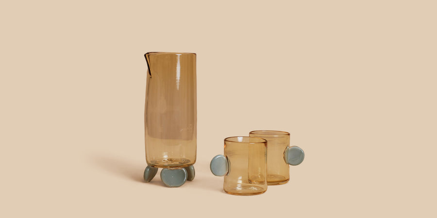 Dot Carafe and Dot Cup Set by Sticky Glass in whiskey and grey, a pale amber carafe standing on blue-grey glass feet with two matching cups
